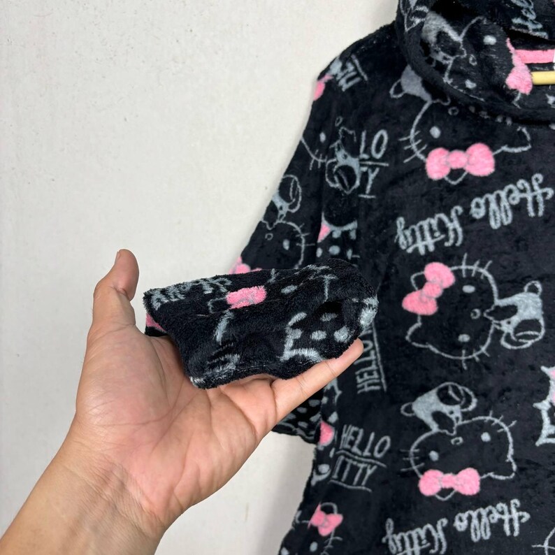 Puede incluir: Sudadera con capucha de felpa negra con un patr&oacute;n repetido de personajes de Hello Kitty, lazos y texto. El dise&ntilde;o incluye detalles en rosa y blanco. La manga est&aacute; sujeta por una mano.