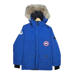 Puede incluir: Una parka de invierno azul brillante con capucha ribeteada de piel. La chaqueta tiene múltiples bolsillos, un cuello negro y un parche con el logotipo circular en el pecho. La parka está diseñada para el clima frío.