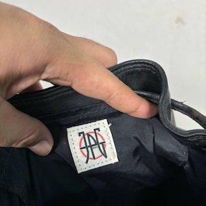 Op de afbeelding: Zwarte leren tas met trekkoord en een wit label met een rood en zwart logo. De binnenkant van de tas is donkerblauw. De tas wordt door een hand vastgehouden.