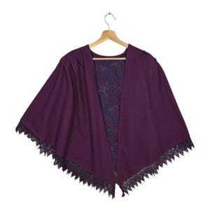 May include: A plum-colored cape with a paisley patterned lining and black lace trim. The cape is hanging on a wooden hanger. The open front design reveals the patterned lining, adding a touch of elegance to the garment.