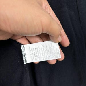 Może przedstawiać: Czarny element odzieży z dołączoną białą metką. Etykieta zawiera tekst w języku angielskim i francuskim, w tym "Made in Japan" i instrukcje pielęgnacji. Skład materiału obejmuje rayon, nylon i angorę.