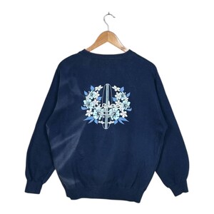 Op de afbeelding: Marineblauw sweatshirt met een bloemen- en surfboard ontwerp. De afbeelding toont een wit surfboard omringd door lichtblauwe en witte bloemen. De sweatshirt heeft lange mouwen en een ronde hals.