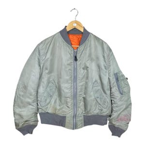 May include: A light green bomber jacket with a gray collar, cuffs, and waistband hangs on a wooden hanger. The jacket has a full zipper closure and two front pockets. The interior lining is bright orange. The jacket appears to be vintage.