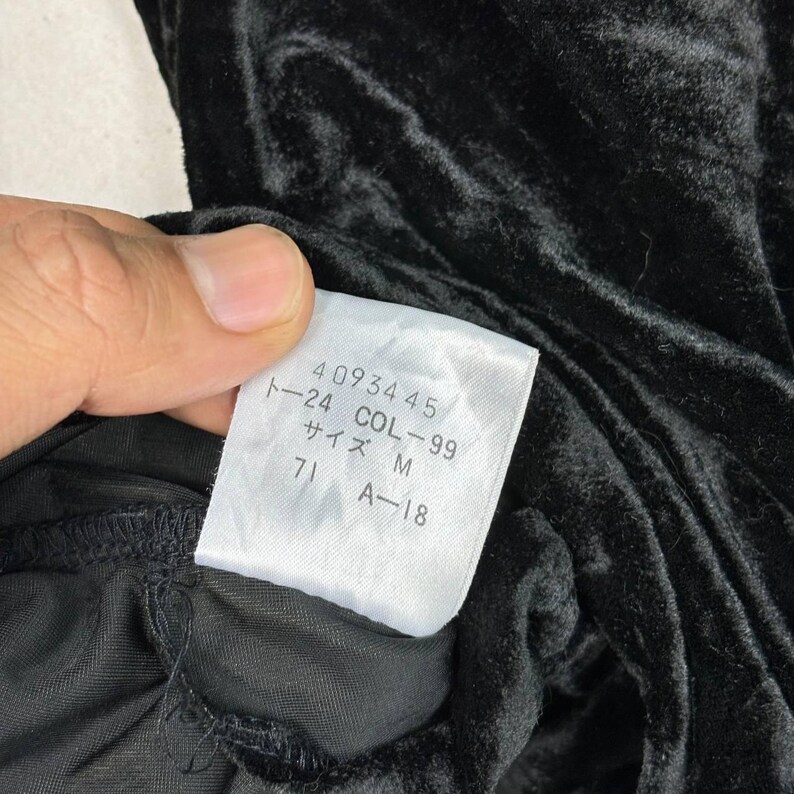 Op de afbeelding: Een wit label met zwarte tekst die "40934 45 COL - 99 24 サイズ M 71 A - 18" luidt, bevestigd aan een zwarte fluwelen stof.