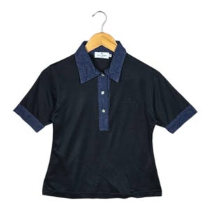 May include: Black polo shirt with a denim collar, placket, and short sleeves. The shirt has a small embroidered logo on the chest and is hanging on a wooden hanger.