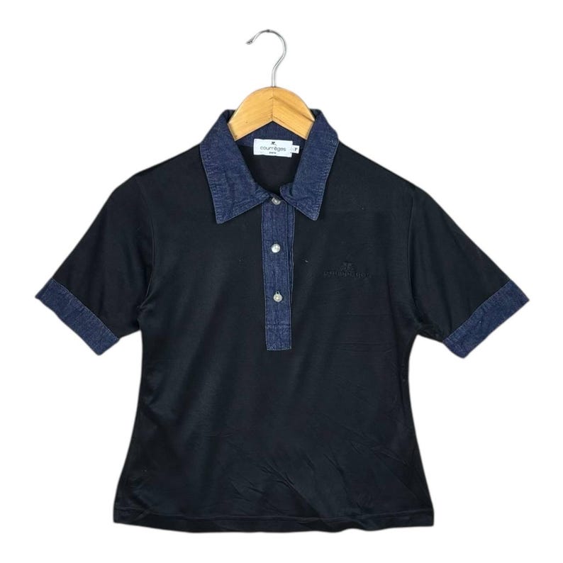 May include: Black polo shirt with a denim collar, placket, and short sleeves. The shirt has a small embroidered logo on the chest and is hanging on a wooden hanger.