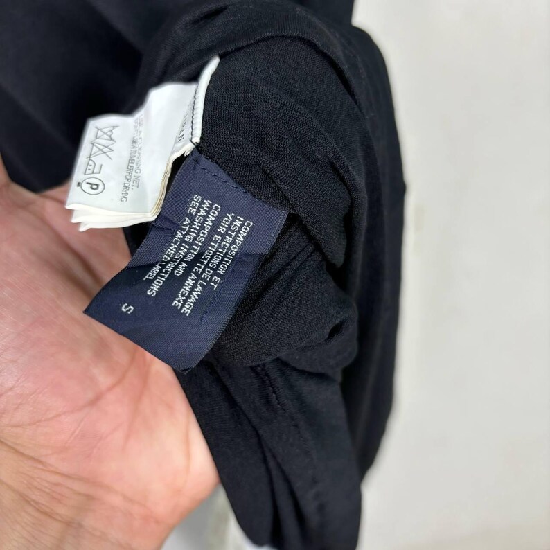 May include: Close-up of a black garment with attached tags. The tags display washing instructions and composition details in both English and French. The garment has a small size label, marked "S".