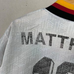 May include: A close-up of a white football shirt with the name "MATTHAUS" printed in large, black letters. The shirt features a geometric pattern and a collar with black, red, and yellow stripes.