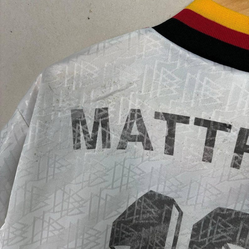 May include: A close-up of a white football shirt with the name "MATTHAUS" printed in large, black letters. The shirt features a geometric pattern and a collar with black, red, and yellow stripes.