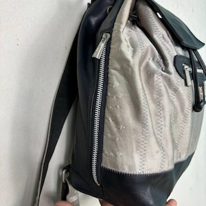 Op de afbeelding: Een vintage rugzak met een combinatie van zwart leer en zilver-grijze stof. De tas heeft een ritssluiting, verstelbare bandjes en een trekkoord aan de bovenkant. Het ontwerp bevat gestikte details en een klepsluiting.