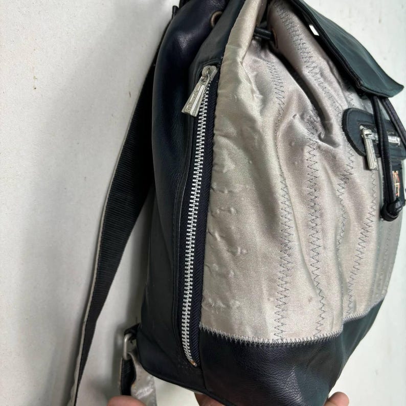 Op de afbeelding: Een vintage rugzak met een combinatie van zwart leer en zilver-grijze stof. De tas heeft een ritssluiting, verstelbare bandjes en een trekkoord aan de bovenkant. Het ontwerp bevat gestikte details en een klepsluiting.