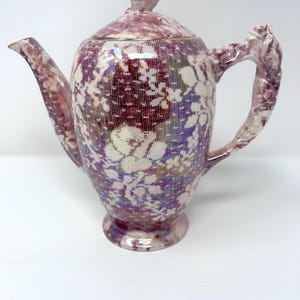 May include: A ceramic teapot featuring a deep purple and white floral lace pattern across its body. The teapot includes a matching lid with a sculpted floral finial and a handle shaped like a textured, organic vine or branch design.