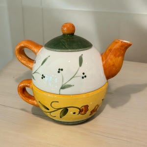May include: A white and yellow ceramic teapot with a green lid and floral design. The teapot has a matching cup.