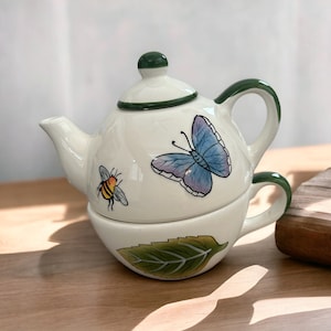 May include: A white ceramic teapot and teacup set with a green handle and lid. The teapot features a blue butterfly and a yellow bee illustration. The teacup has a green leaf design.