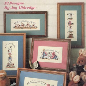 May include: A cross-stitch pattern booklet titled "Make a Joyful Noise" with 12 designs by Joy Alldredge. The cover features a white brick background with framed cross-stitch patterns and plush bunny and bear toys.