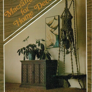 May include: A brown wooden cabinet with two doors and a macrame hanging plant holder with a brown rope. The macrame hanging plant holder is hanging from the ceiling and has a plant in it. The cabinet is sitting on a brown carpet. The text "Macrame for Home Decor" is in the top left corner of the image.