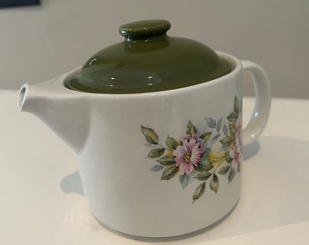 May include: A white ceramic teapot with a green lid and floral design. The teapot has a handle and a spout. The floral design features pink and yellow flowers with green leaves.