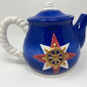 May include: A blue ceramic teapot with a white rope handle and a white star with a gold steering wheel design. The teapot has a white lid with a rope detail.