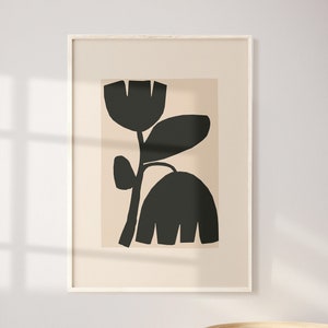May include: Framed abstract art print featuring a black floral design on a beige background. The artwork is displayed in a light-colored wooden frame. A wooden bowl and decorative spheres sit on a table in front of the artwork.