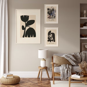 May include: Three framed art prints with abstract designs hang on a neutral wall. The largest print features a black floral silhouette on a beige background. The other two prints display abstract shapes and text. A lamp and furniture are in the foreground.