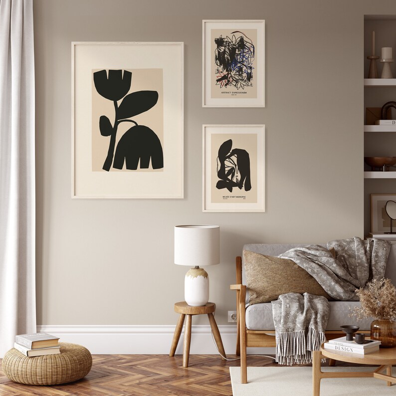 May include: Three framed art prints with abstract designs hang on a neutral wall. The largest print features a black floral silhouette on a beige background. The other two prints display abstract shapes and text. A lamp and furniture are in the foreground.