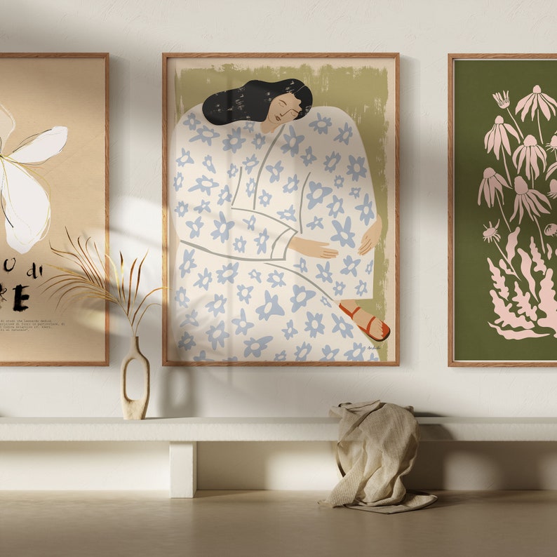 May include: Three framed illustrations with a muted color palette. The center illustration features a woman in a white dress with a blue floral pattern, sitting with her eyes closed. The left illustration features a white flower with a gold stem on a beige background. The right illustration features pink flowers and leaves on a green background.