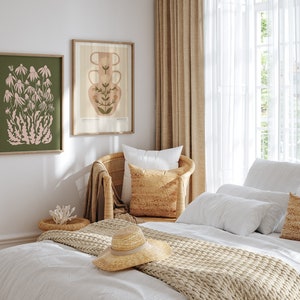 May include: A bedroom with a white bed, a wicker chair, and two framed botanical prints on the wall. The bed has a white duvet, a beige knitted throw, and a straw hat on top. The prints feature a pink flower and a vase with flowers.