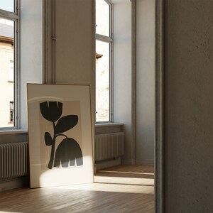 May include: A framed art print featuring a stylized black floral design against a beige background. The artwork is displayed in a room with natural light, leaning against a wall near a window. The frame is a light brown color.