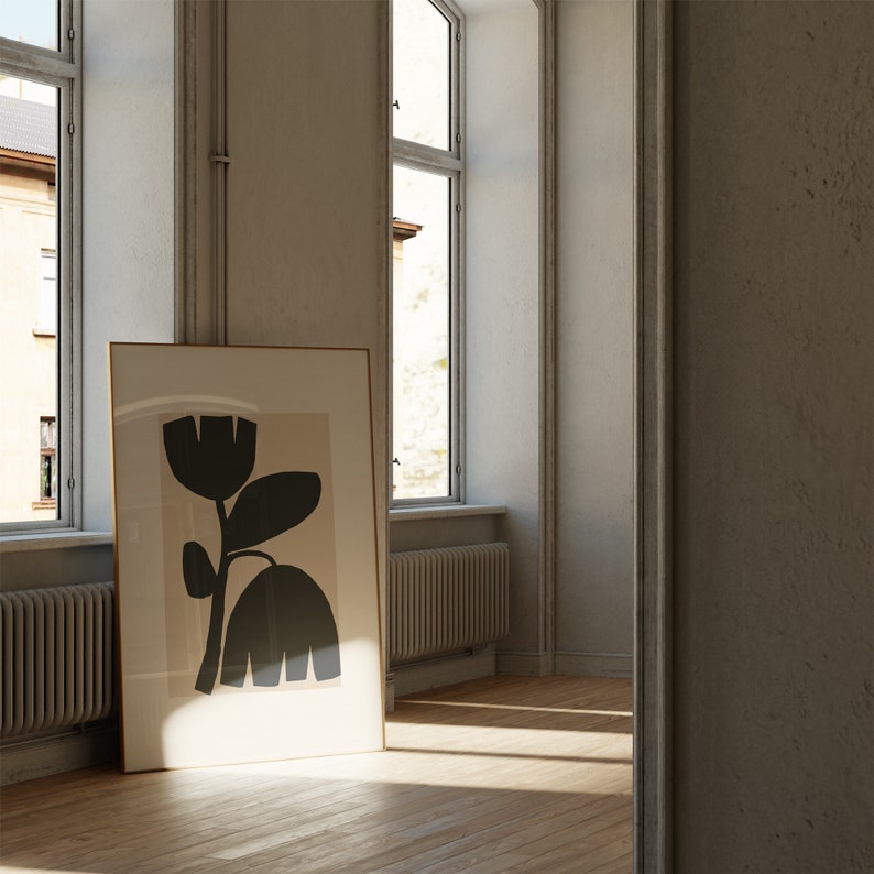 May include: A framed art print featuring a stylized black floral design against a beige background. The artwork is displayed in a room with natural light, leaning against a wall near a window. The frame is a light brown color.
