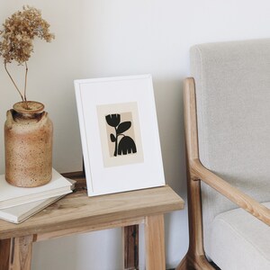 May include: A framed art print featuring a black floral design on a beige background. The artwork is displayed on a wooden side table next to a beige armchair. A rustic brown vase with dried flowers sits on the table.