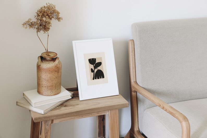 May include: A framed art print featuring a black floral design on a beige background. The artwork is displayed on a wooden side table next to a beige armchair. A rustic brown vase with dried flowers sits on the table.