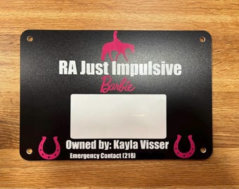 以下が含まれることがあります： 黒色の馬小屋の看板に、白い文字で「RA Just Impulsive Barbie」と「Owned by: Kayla Visser」と書かれており、両側にピンクの馬蹄が描かれています。看板には、名前タグ用の白い長方形もあります。