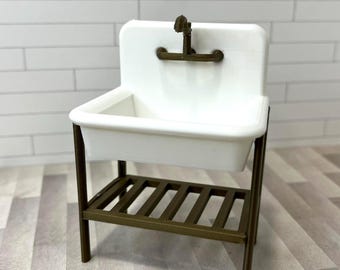 Op de afbeelding: Een miniatuur wit porseleinen wastafel met een bronzen kraan en frame. De wastafel heeft een rechthoekige bak en een plank met latten eronder. Het frame en de kraan hebben een vintage esthetiek.