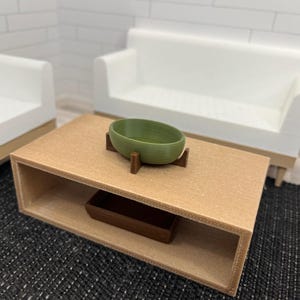 May include: A green ceramic bowl on a wooden stand sits on a light brown coffee table with a shelf. The table is in front of two white couches.
