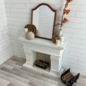 May include: A white fireplace mantel with a brown wooden mirror above it. The mantel has a white fireplace with a black grate and a brick hearth. There are two white candles, a brown vase with dried flowers, and a brown ceramic vase with a wooden tray on the mantel. There is a black metal log holder with logs on the floor.