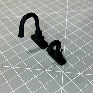 May include: Two black, 3D-printed, hook-shaped objects. Each has a rectangular base and a curved, arched hook extending upwards. The objects are positioned on a gray grid surface, with one casting a shadow.