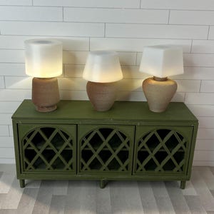 Könnte beinhalten: Drei Miniatur-Lampen mit weißen Schirmen und strukturierten braunen Sockeln stehen auf einem grünen Schrank. Der Schrank hat ein Gittermuster auf den Türen. Die Lampen sind beleuchtet und geben ein warmes Licht ab.