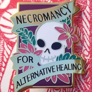 Puede incluir: Un pin de esmalte dorado con fondo morado. El pin presenta una calavera blanca con ojos negros y una flor rosa. El texto "NECROMANCY FOR ALTERNATIVE HEALING" está escrito en una pancarta dorada.