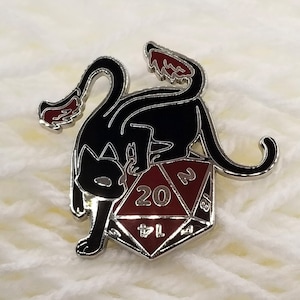 Könnte beinhalten: Eine schwarze Emaille-Anstecknadel mit einer Katze mit roten Flammen am Schwanz, die auf einem roten 20-seitigen Würfel sitzt.