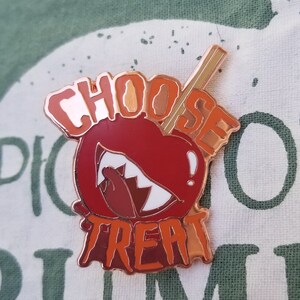 Pode incluir: Um broche de esmalte vermelho e laranja com um design de ma&ccedil;&atilde; de caramelo. A ma&ccedil;&atilde; tem um sorriso branco e dentado e um palito que sai do topo. O texto "Choose Treat" est&aacute; escrito em uma fonte que pinga ao redor da ma&ccedil;&atilde;.