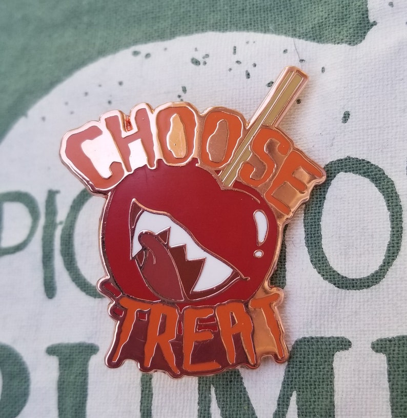 Pode incluir: Um broche de esmalte vermelho e laranja com um design de ma&ccedil;&atilde; de caramelo. A ma&ccedil;&atilde; tem um sorriso branco e dentado e um palito que sai do topo. O texto "Choose Treat" est&aacute; escrito em uma fonte que pinga ao redor da ma&ccedil;&atilde;.