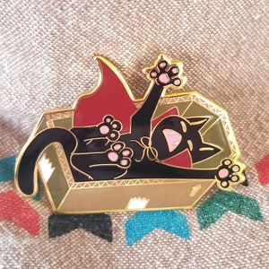 May include: Gold-tone enamel pin featuring a black cat wearing a red cape and lying in a coffin. The cat has pink paws and a wide open mouth. The coffin has a number "1" on the side.