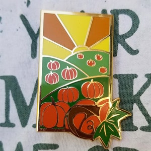 Puede incluir: Pin de esmalte dorado con una puesta de sol sobre un campo de calabazas. El pin es verde, naranja, amarillo y marrón. Las calabazas son naranjas y las hojas son verdes.
