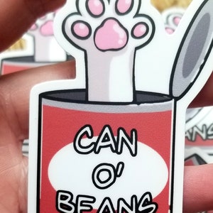 May include: A white paw print sticker with pink paw pads inside a red can with the text "CAN O' BEANS" on a white background.