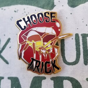 Pode incluir: Pin de esmalte dourado com uma aranha vermelha, branca e marrom com as palavras "Choose Trick" em letras pretas.