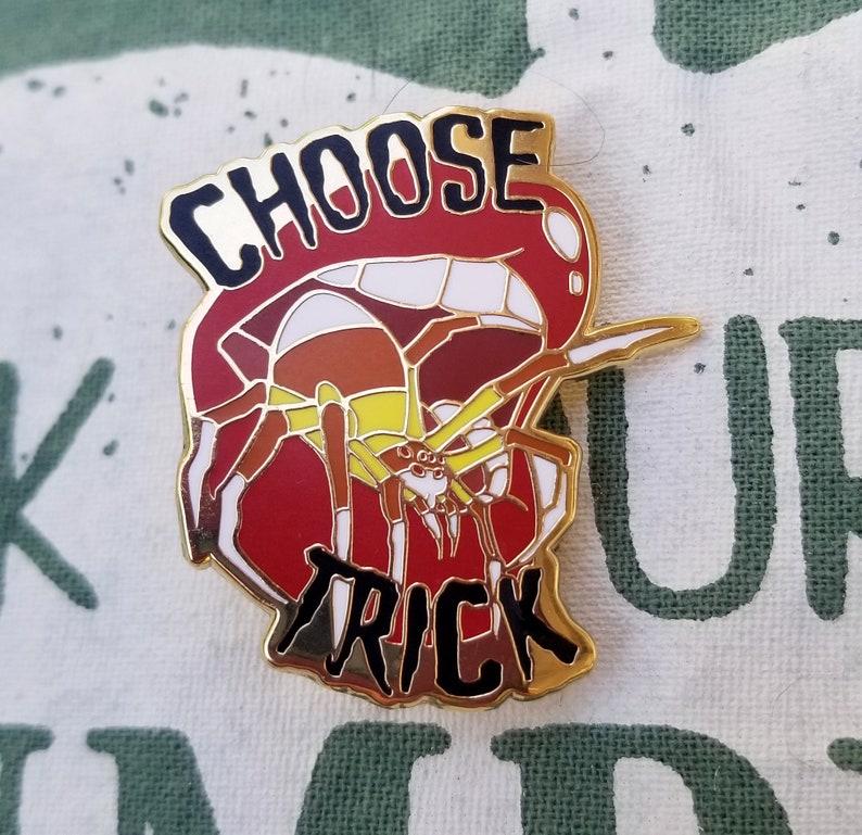 Pode incluir: Pin de esmalte dourado com uma aranha vermelha, branca e marrom com as palavras "Choose Trick" em letras pretas.