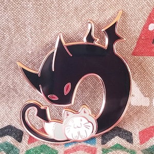May include: A black and gold enamel pin featuring a black cat curled around a smaller white cat with pink ears and nose. The black cat has a red eye visible.