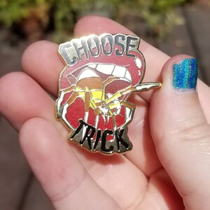 Pode incluir: Pin dourado com um design vermelho, branco e preto. O pin apresenta uma aranha com um abd&ocirc;men amarelo e as palavras "Choose Trick" em preto.