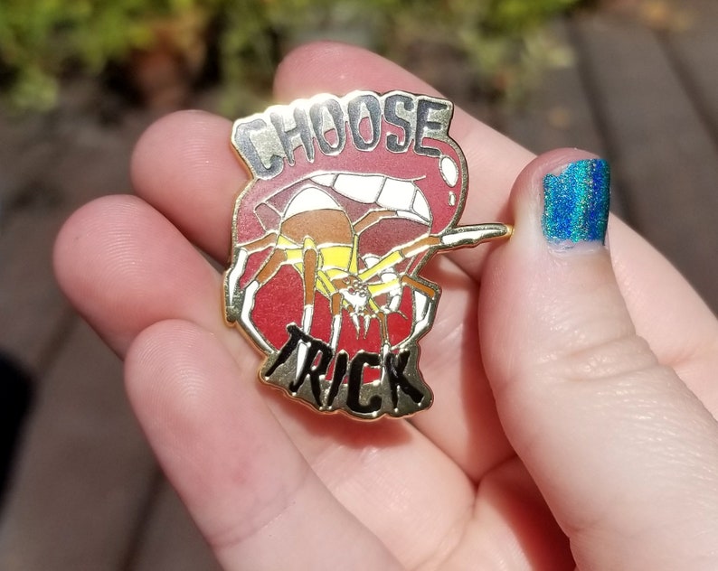 Pode incluir: Pin dourado com um design vermelho, branco e preto. O pin apresenta uma aranha com um abd&ocirc;men amarelo e as palavras "Choose Trick" em preto.