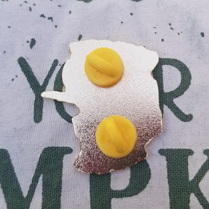 Pode incluir: O verso de um alfinete de metal dourado com um design amarelo. O alfinete tem dois fechos de metal amarelo na parte de tr&aacute;s.